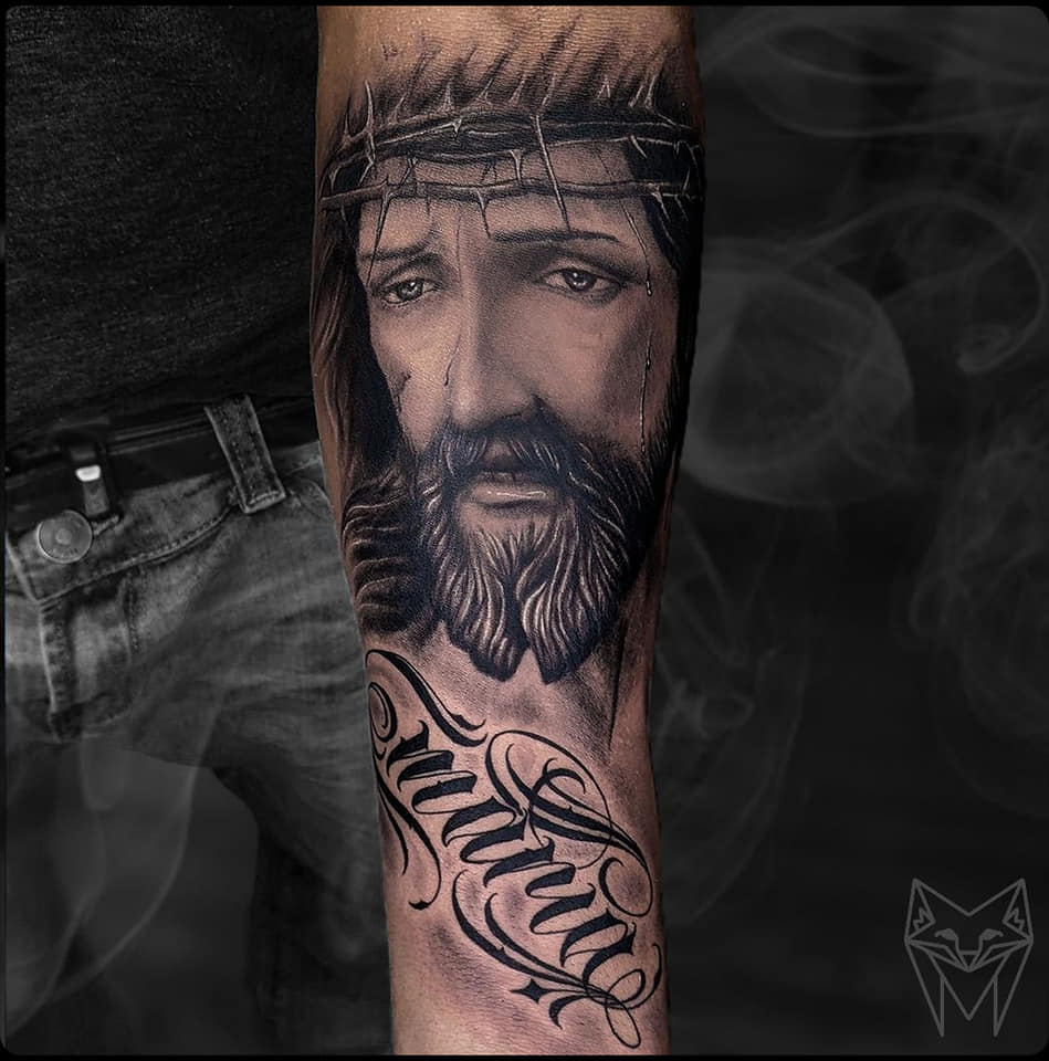 Black and grey realism tattoo of a Jesus portrait with a crown of thorns above chicano script, on a forearm